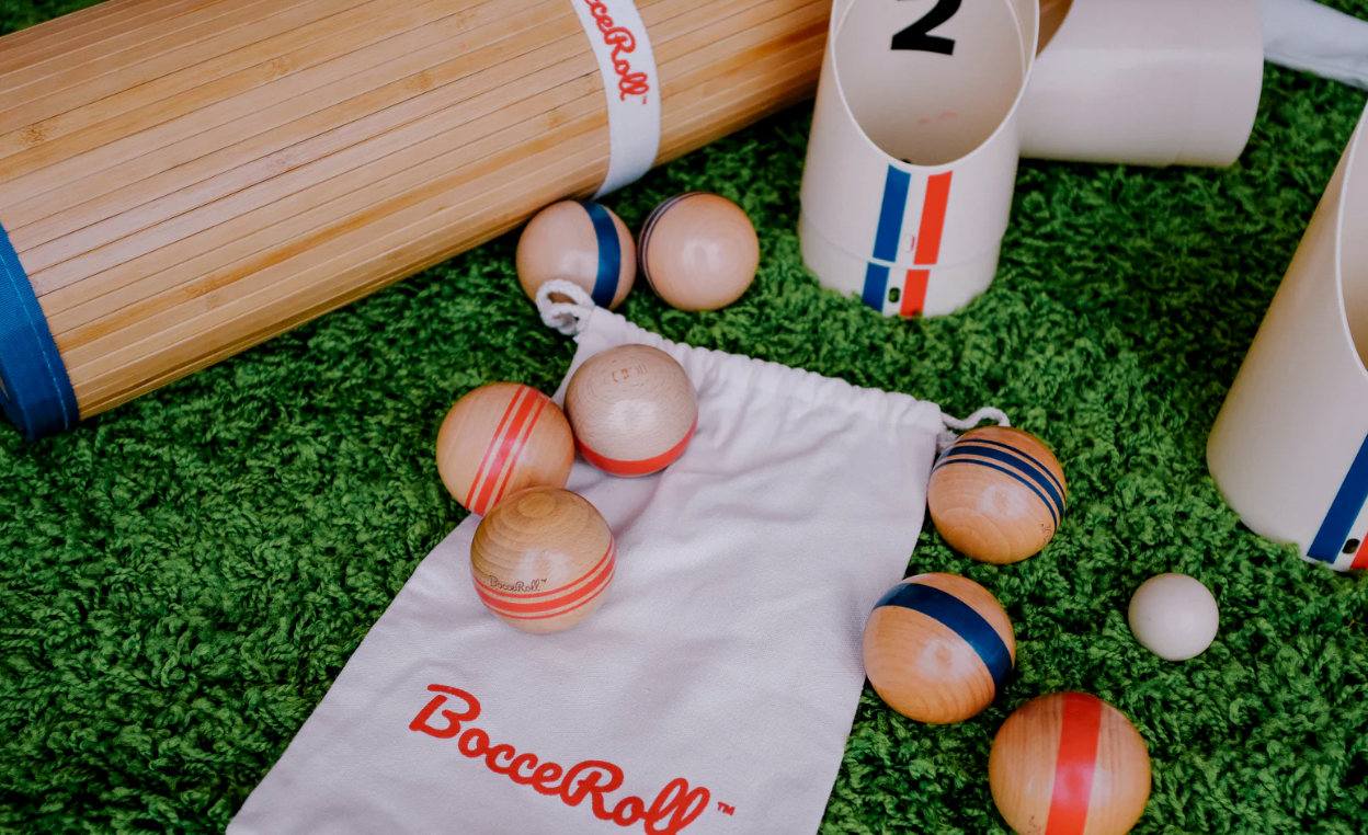 Bocce ball set with wooden balls, white cups, and a drawstring bag on grass.